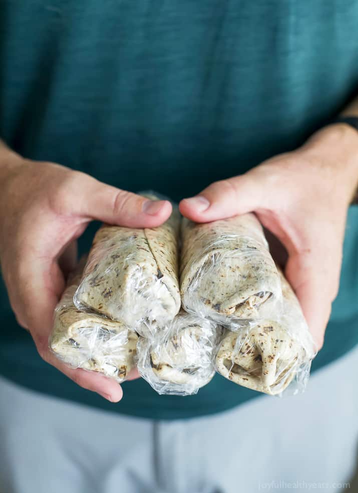 Hands holding several Freezer Bacon & Egg Breakfast Burritos wrapped in plastic wrap