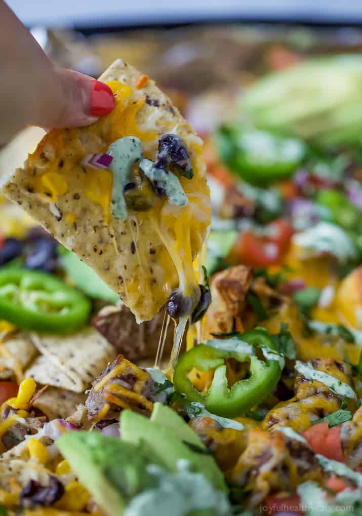 A hand lifting a tortilla chip from steak nachos on a sheet pan.