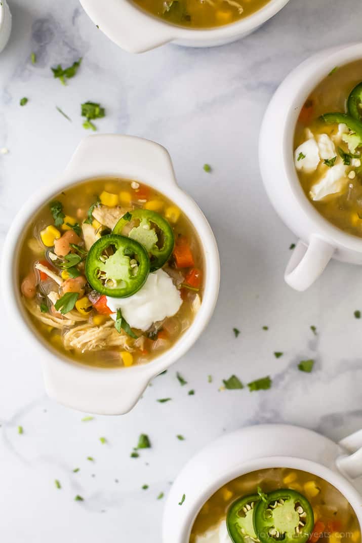 This healthy white chicken chili recipe is filled with juicy chicken, hearty pinto beans, tender corn, and more. Perfect for chilly days! Top view of crocks of white chicken chili with jalapeno slices and sour cream.