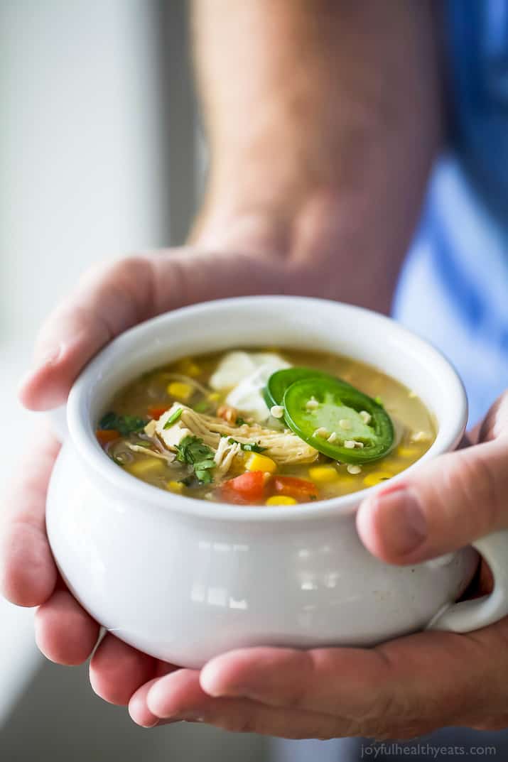 This healthy white chicken chili recipe is filled with juicy chicken, hearty pinto beans, tender corn, and more. Perfect for chilly days! Hands holding a bowl of white chicken chili with jalapeno slices and sour cream.