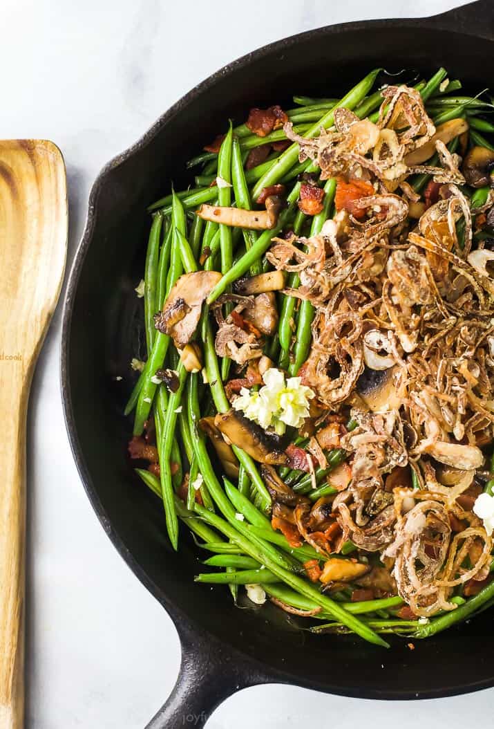 Green Bean Casserole is made with sautéed green beans, savory mushrooms and crispy bacon topped with homemade fried shallots. Healthy green bean casserole in a skillet near a wooden spoon.