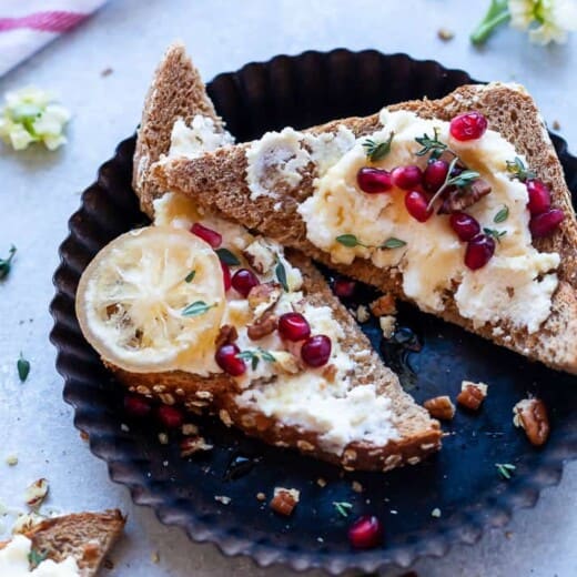 Sliced ricotta cheese toast with fresh pomegranate, candied pecans, thyme, and honey on top.