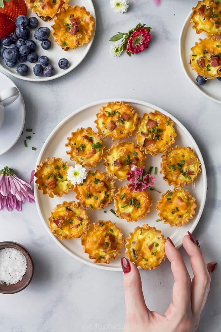 ham and cheese mini quiches topped with chives on a plate with a hand grabbing one of the quiches