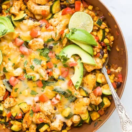 Overhead view of a chicken enchilada skillet topped with melted cheese, avocado slices, and lime.