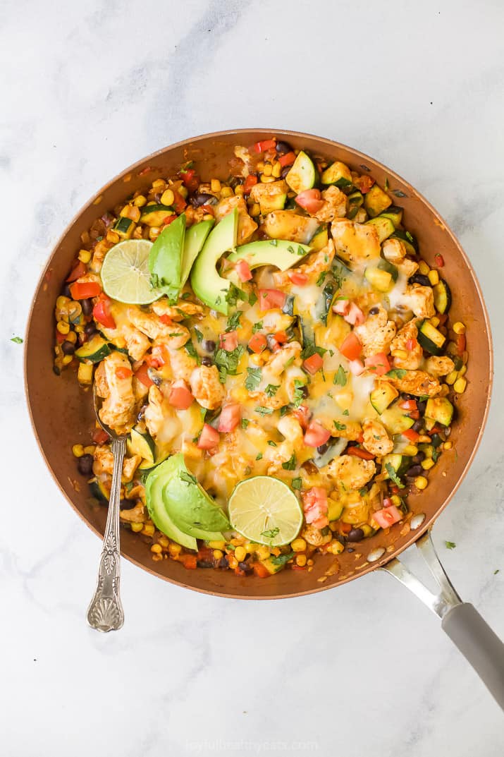 Overhead view of a chicken enchilada skillet topped with melted cheese, avocado slices, and lime.