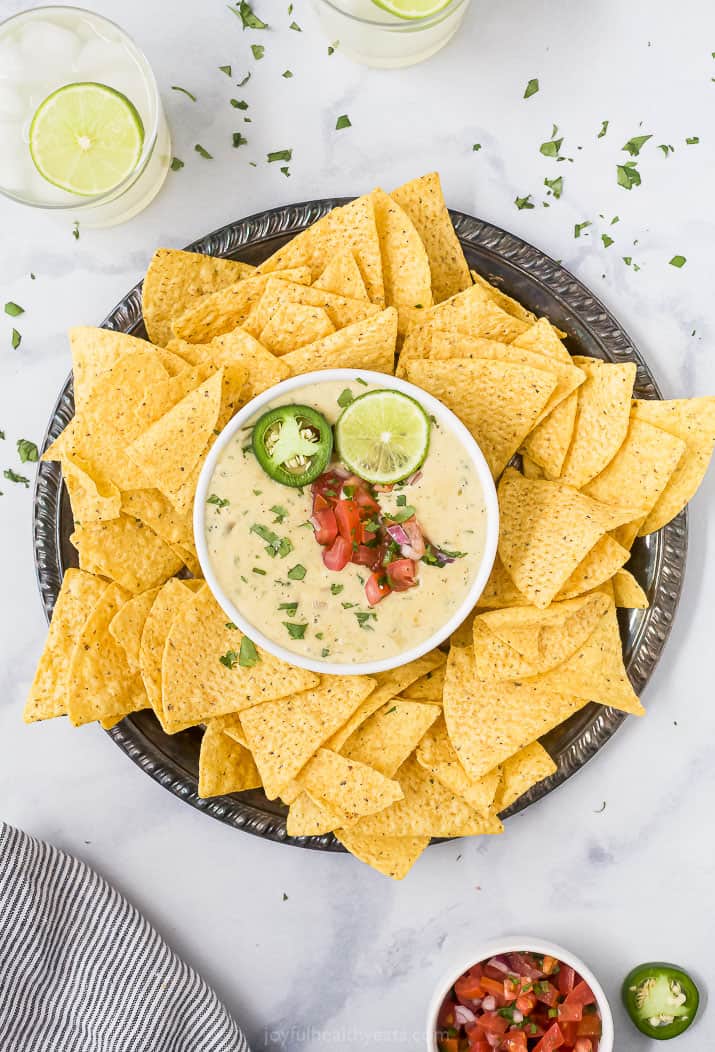 Overhead view of a bowl of white queso dip garnished with lime and pico de gallo in the center of a plate surrounded by tortilla chips.