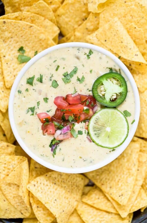Overhead view of a bowl of white queso dip garnished with lime and pico de gallo surrounded by tortilla chips.