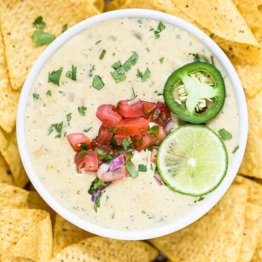 Overhead view of a bowl of white queso dip garnished with lime and pico de gallo surrounded by tortilla chips.