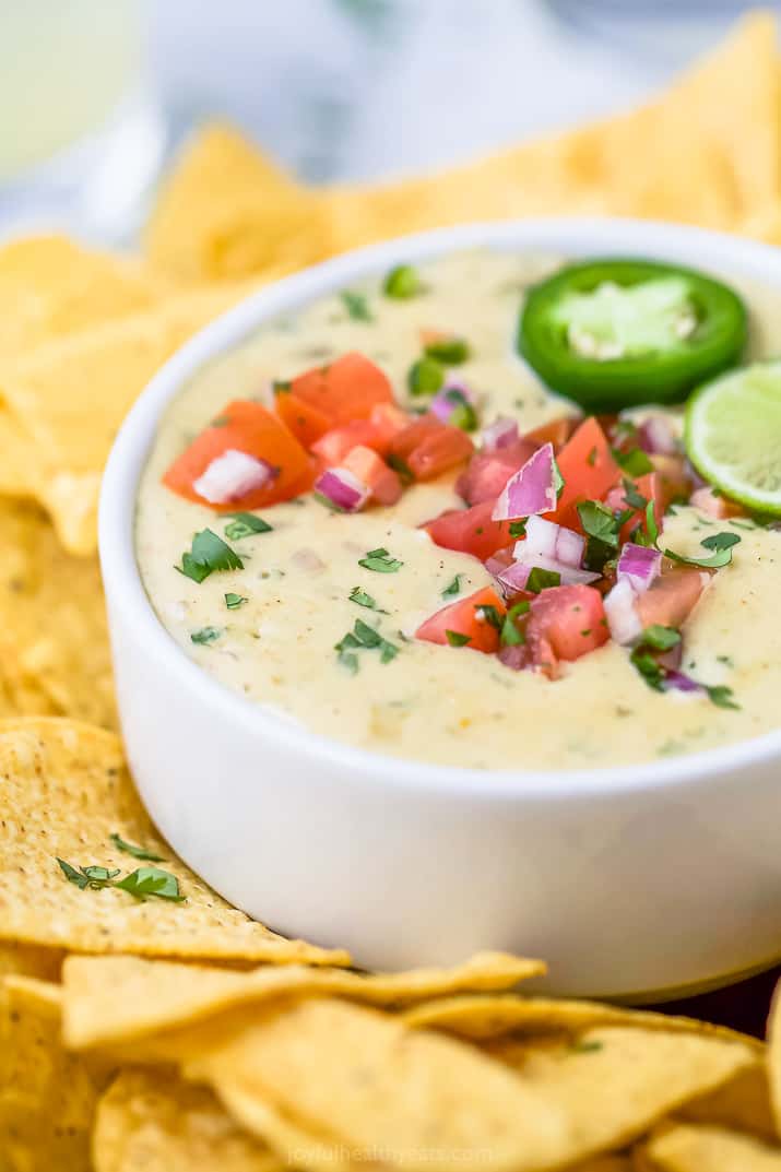 A bowl of white queso dip garnished with lime and pico de gallo surrounded by tortilla chips.