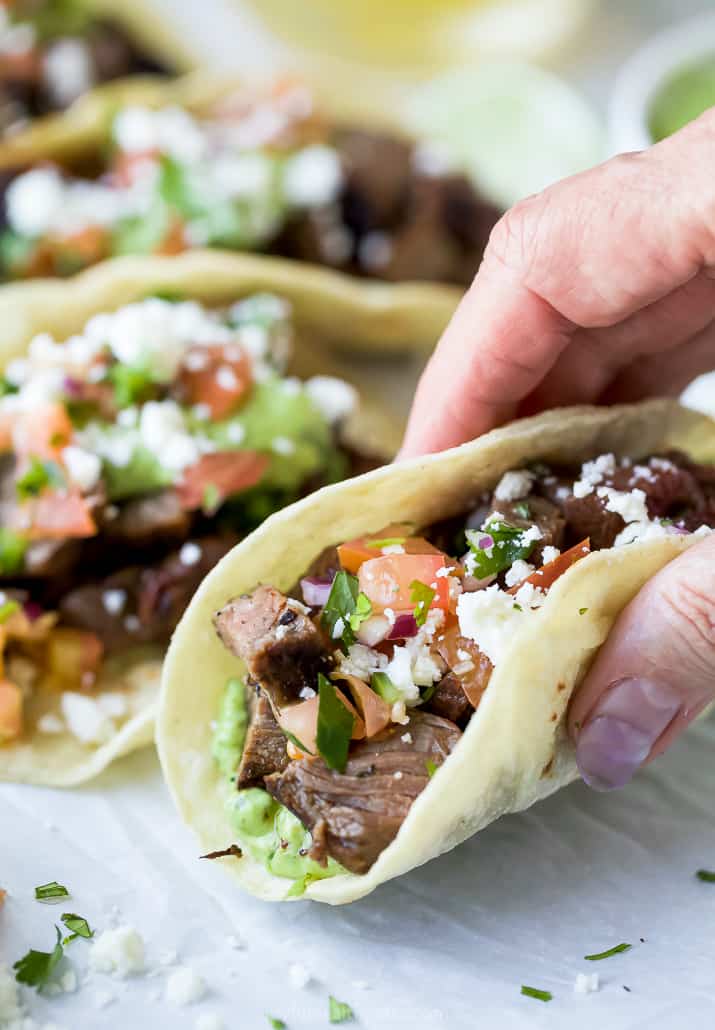 a hand holding a corn tortilla filled with carne asada taco meat and avocado crema