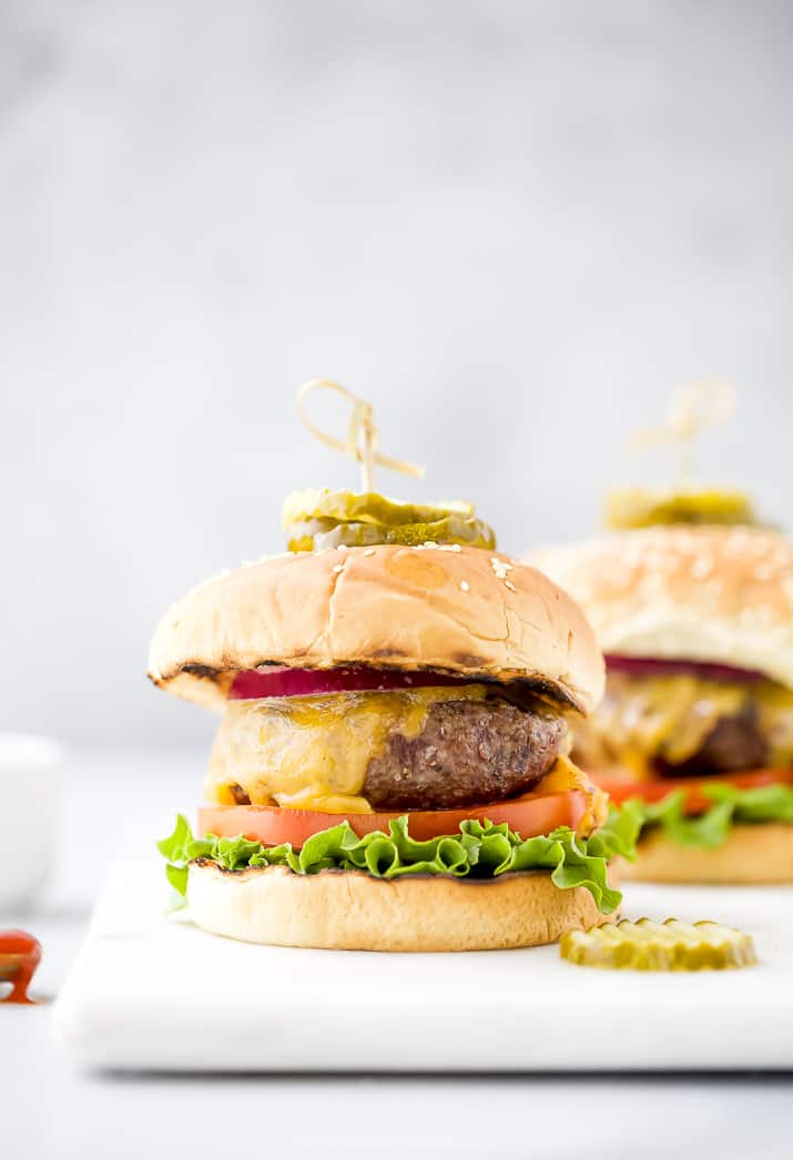 grilled burgers topped with cheese, lettuce, tomato, onion, and pickles sitting on a white cutting board