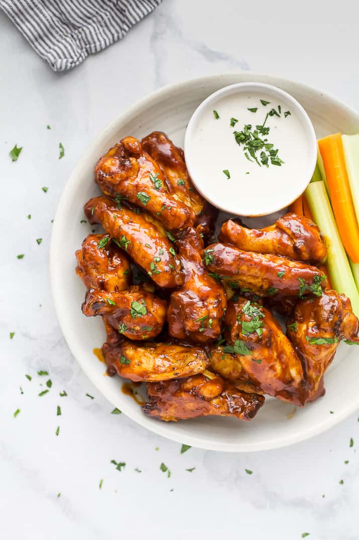 Make extra crispy air fryer chicken wings smothered in a sweet and sticky homemade honey BBQ sauce. Game day appetizers just got better! Close up overhead view of air fryer chicken wings on a white plate next to a side of crudités and ranch dressing.
