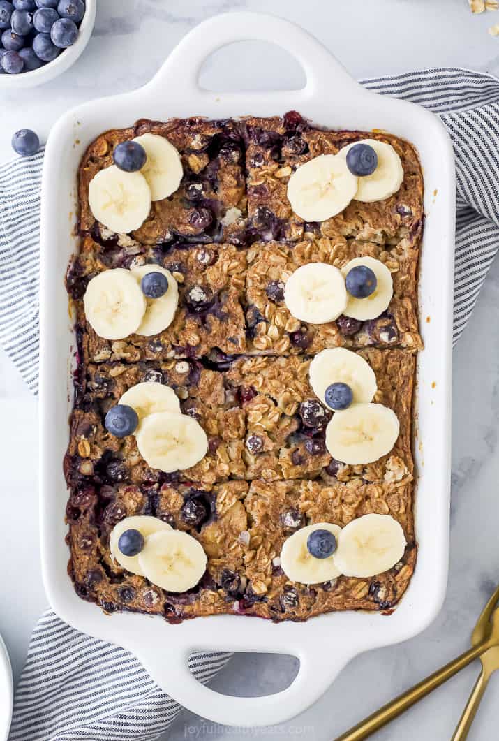 A white baking dish of blueberry baked oatmeal topped with slices of banana and fresh blueberries