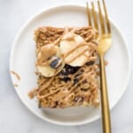 A slice of baked oatmeal on a white plate with a gold fork, topped with fresh blueberries, banana slices and almond butter