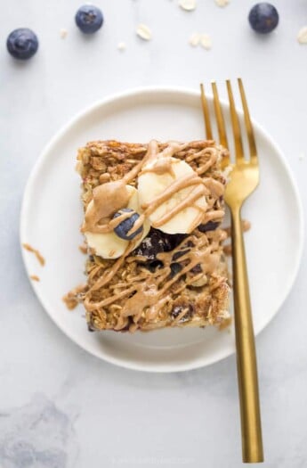 A slice of baked oatmeal on a white plate with a gold fork, topped with fresh blueberries, banana slices and almond butter