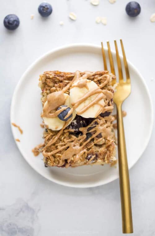A slice of baked banana blueberry oatmeal on a white plate with a gold fork, topped with fresh blueberries, banana slices and almond butter