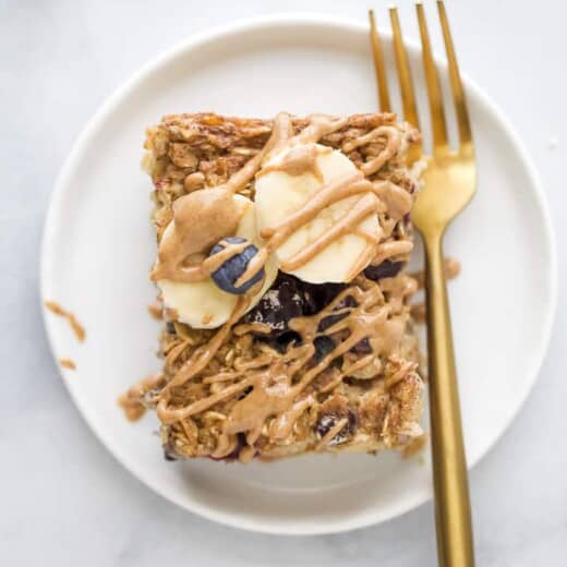 A slice of baked oatmeal on a white plate with a gold fork, topped with fresh blueberries, banana slices and almond butter