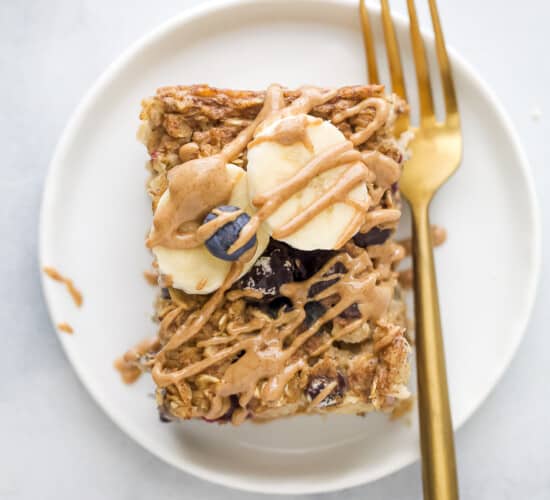 A slice of baked oatmeal on a white plate with a gold fork, topped with fresh blueberries, banana slices and almond butter