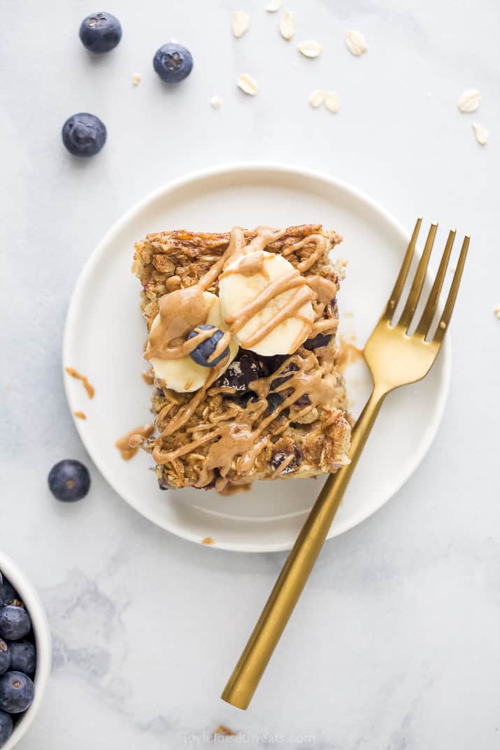 Baked oatmeal topped with banana slices, blueberries and almond butter on a small white plate with a gold fork.