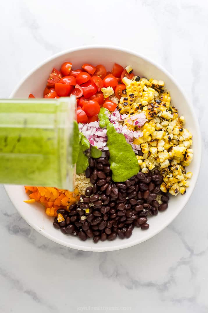 A big white bowl with tomato, charred corn, black beans, pepper, quinoa and onion, being topped with cilantro dressing to make a Mexican Quinoa Salad.