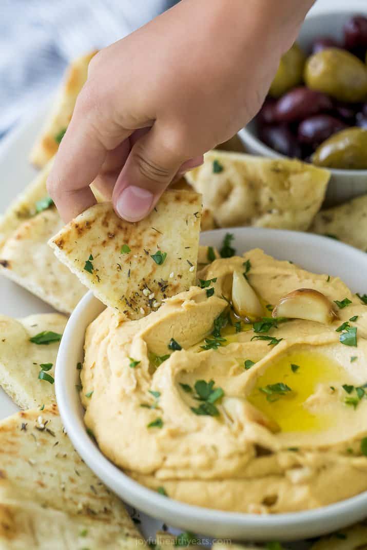 A bowl of roasted garlic hummus surrounded by pita chips and olives