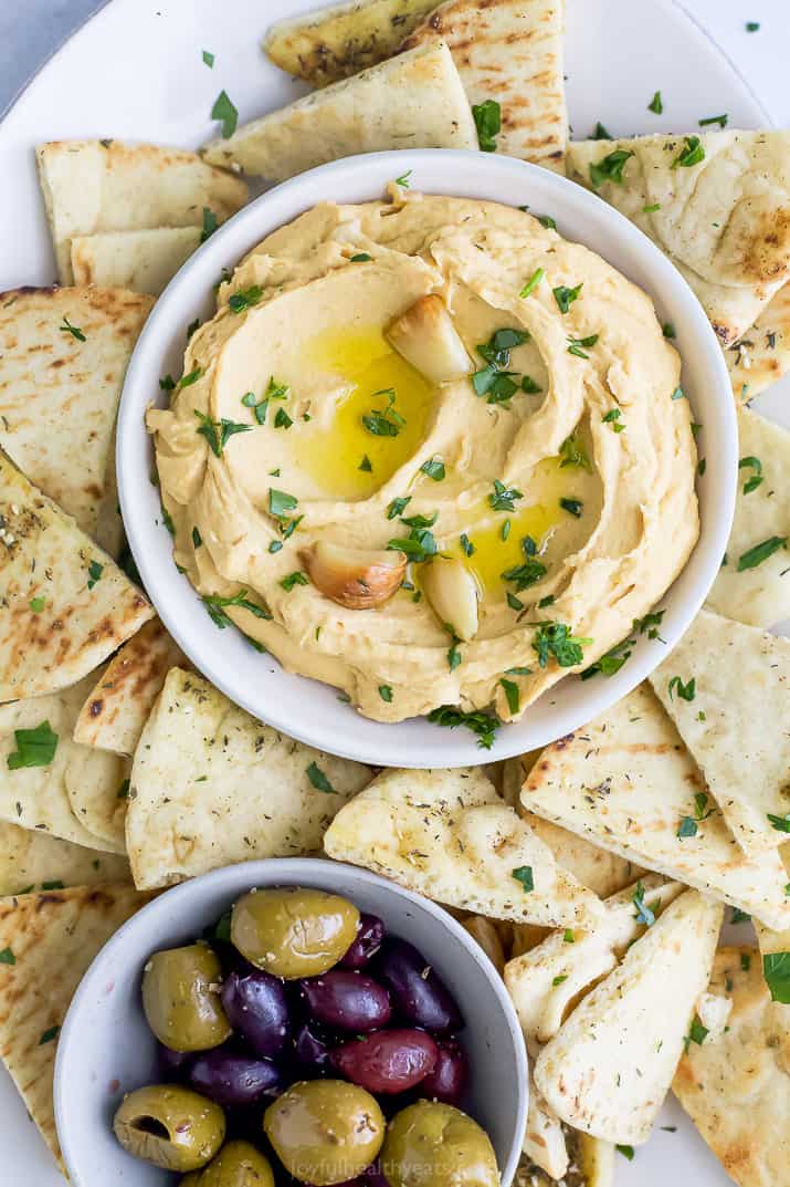 A bowl of roasted garlic hummus surrounded by pita chips and olives