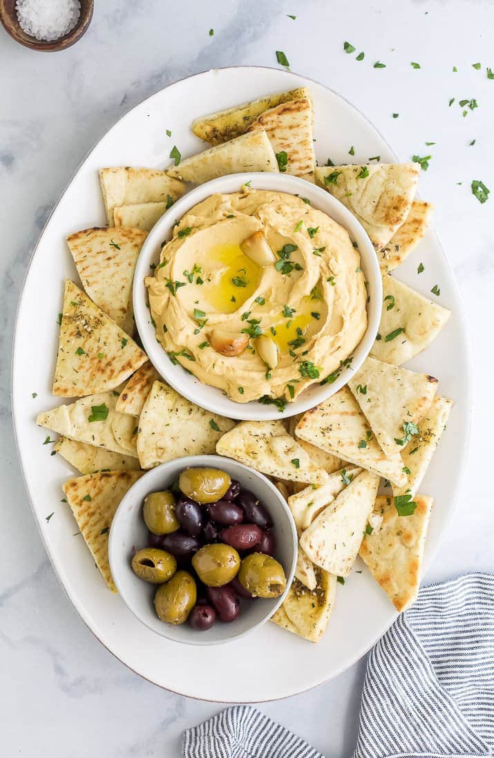 A bowl of roasted garlic hummus surrounded by pita chips and olives