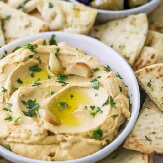 A bowl of roasted garlic hummus surrounded by pita chips and olives