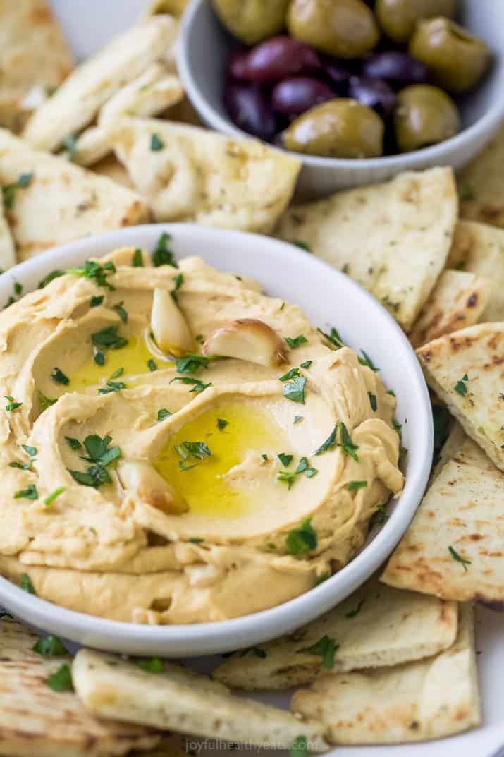 A bowl of roasted garlic hummus surrounded by pita chips and olives