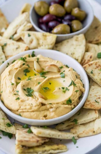 A bowl of roasted garlic hummus surrounded by pita chips and olives