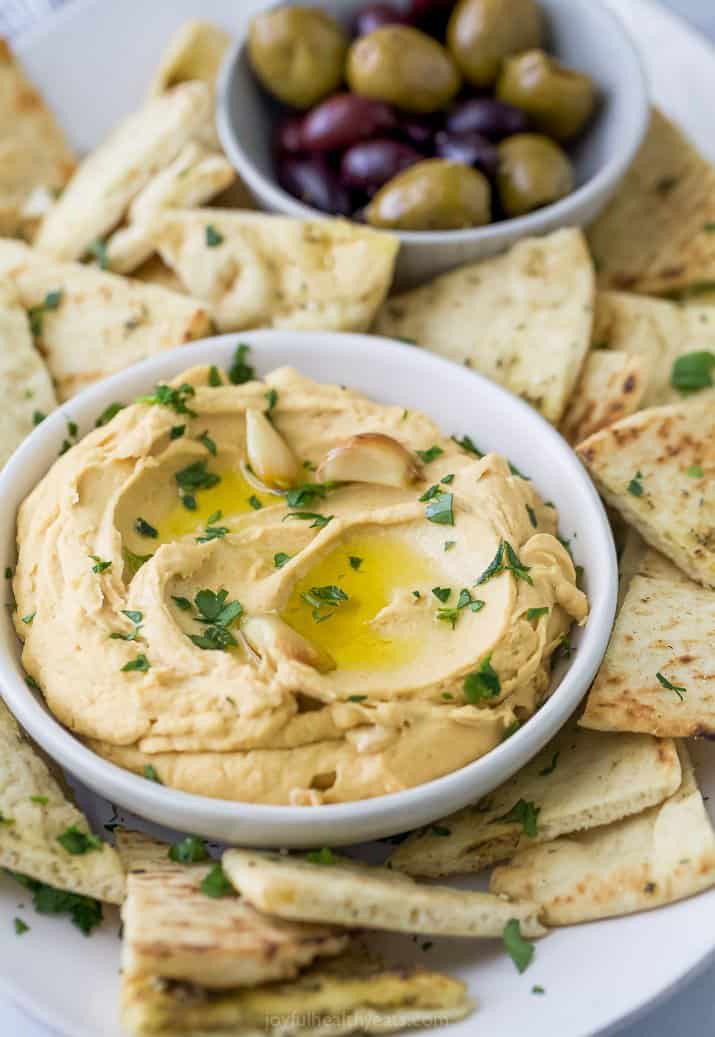 A bowl of roasted garlic hummus surrounded by pita chips and olives