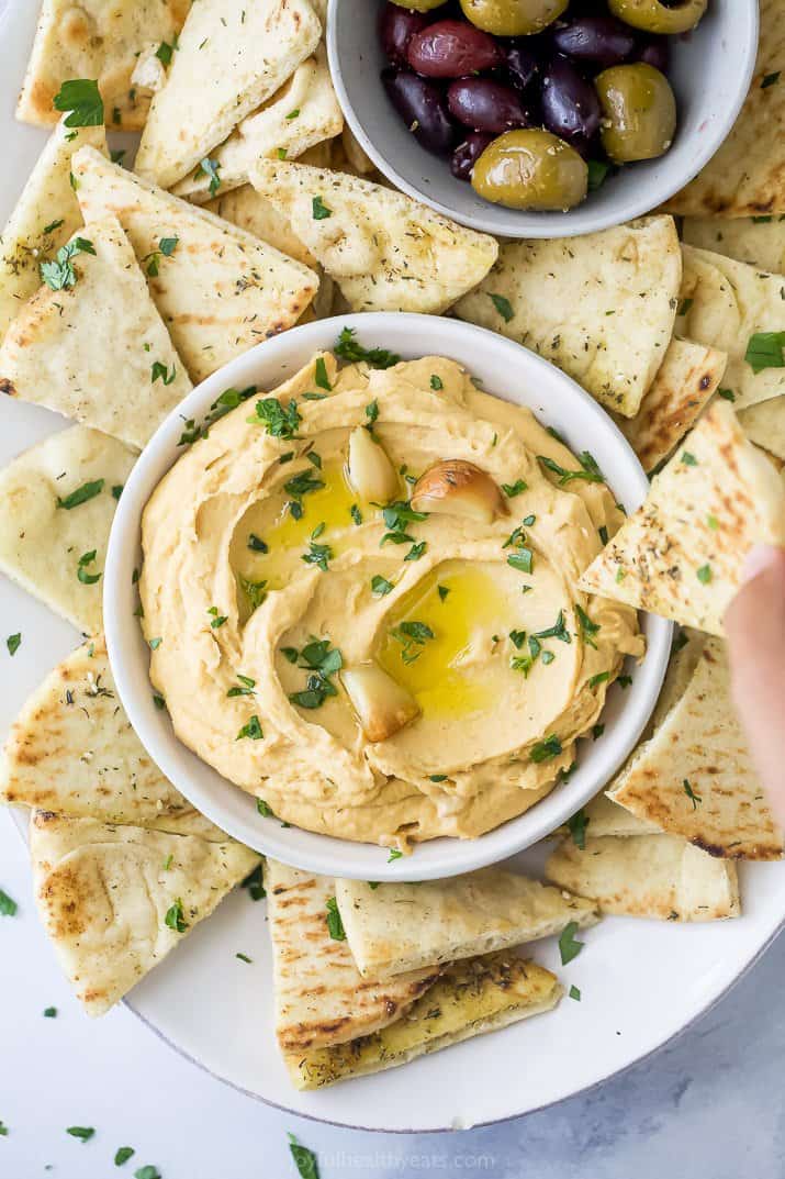 A bowl of roasted garlic hummus surrounded by pita chips and olives