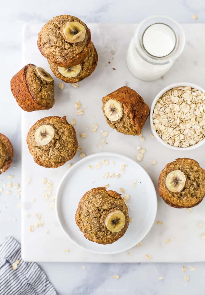 Aerial view of a banana muffin on a plate with more muffins, milk, and a bowl of oats next to it