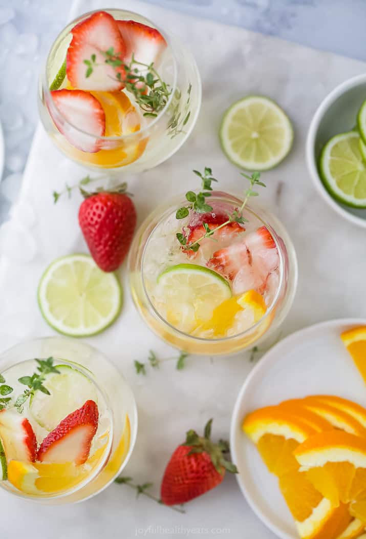 An aerial view of three glasses of white wine spritzer, with orange and lemon slices and strawberry