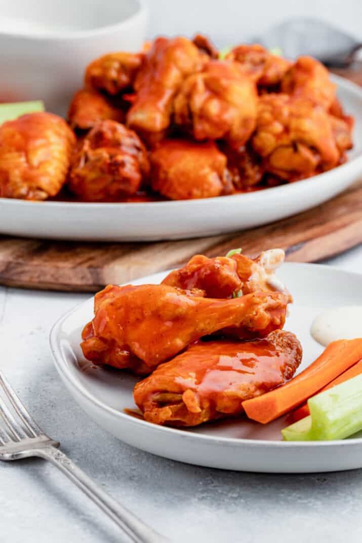 A Serving of Baked Buffalo Chicken Wings on a White Plate with a Platter Full of Wings in the Background