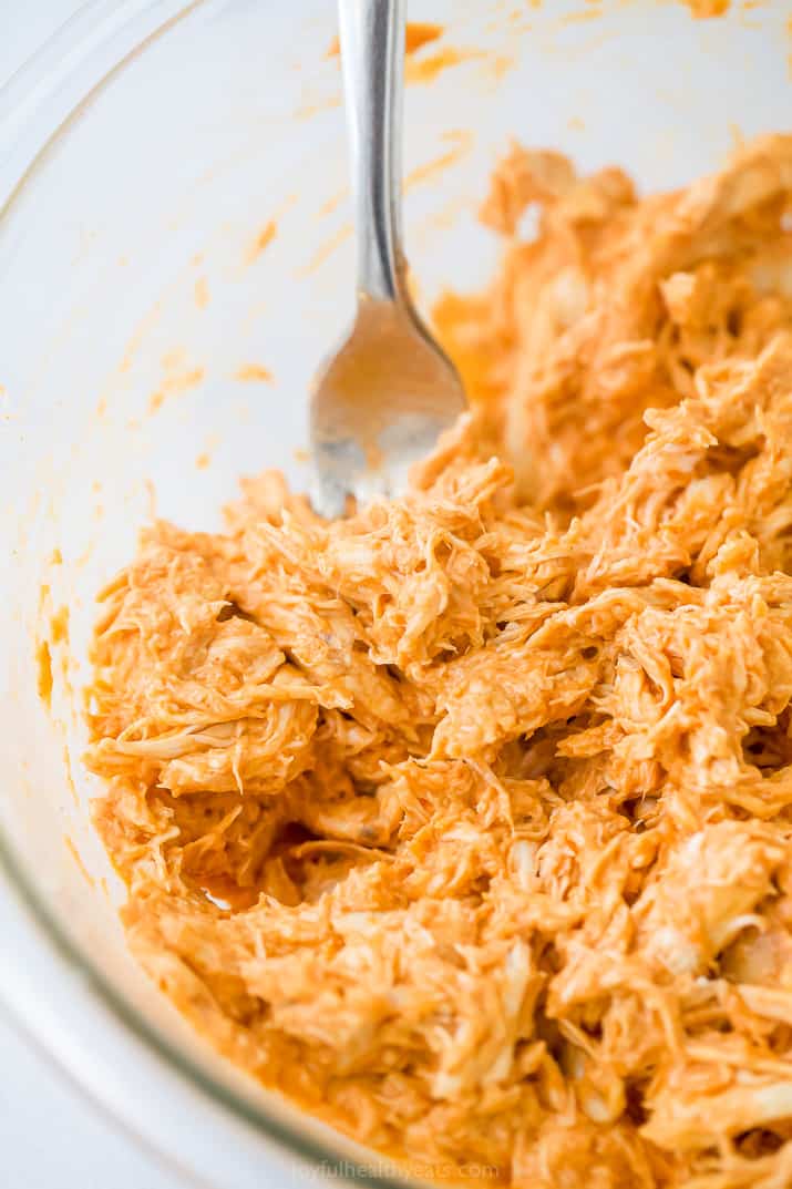 The shredded buffalo chicken mixture in a glass mixing bowl with a metal fork