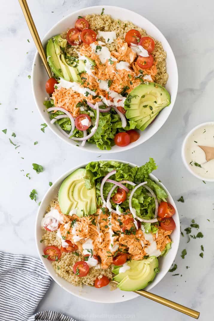 Two buffalo chicken quinoa bowls on a gray and white counter beside a striped kitchen towel