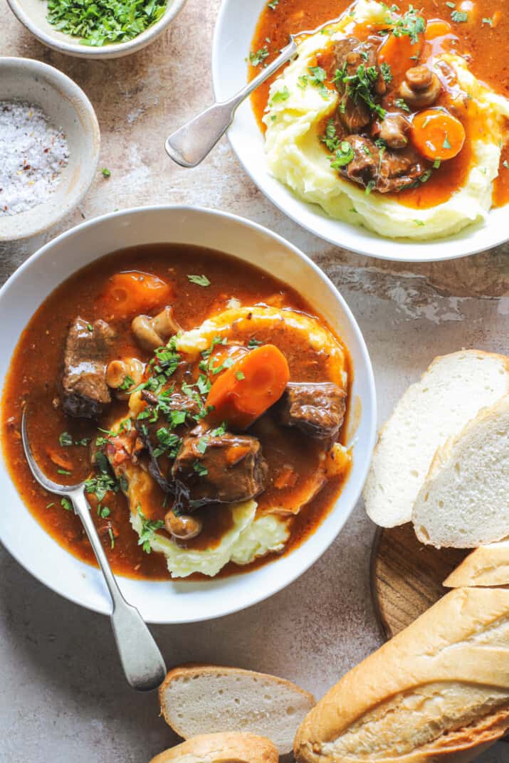 Two bowls of slow cooker beef bourguignon beside a loaf and slices of French bread