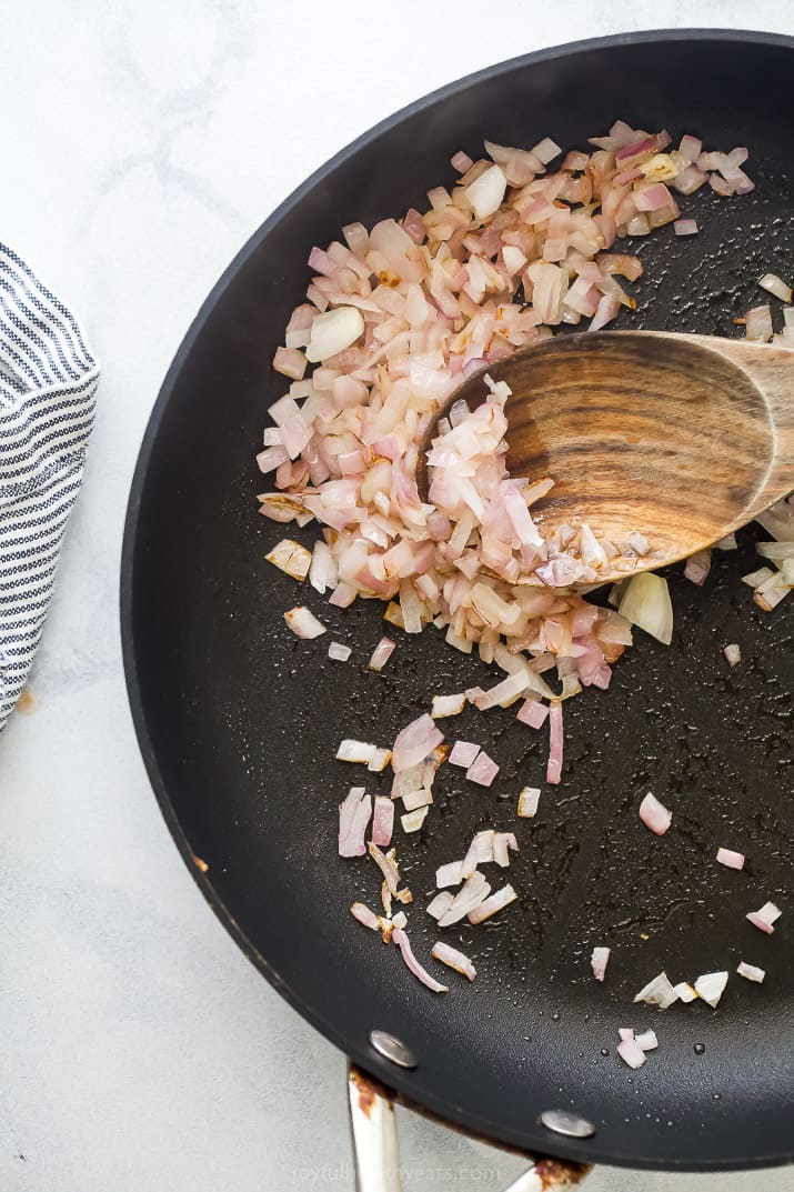 Diced red onions and garlic being sautéd with a striped kitchen towel beside the skillet