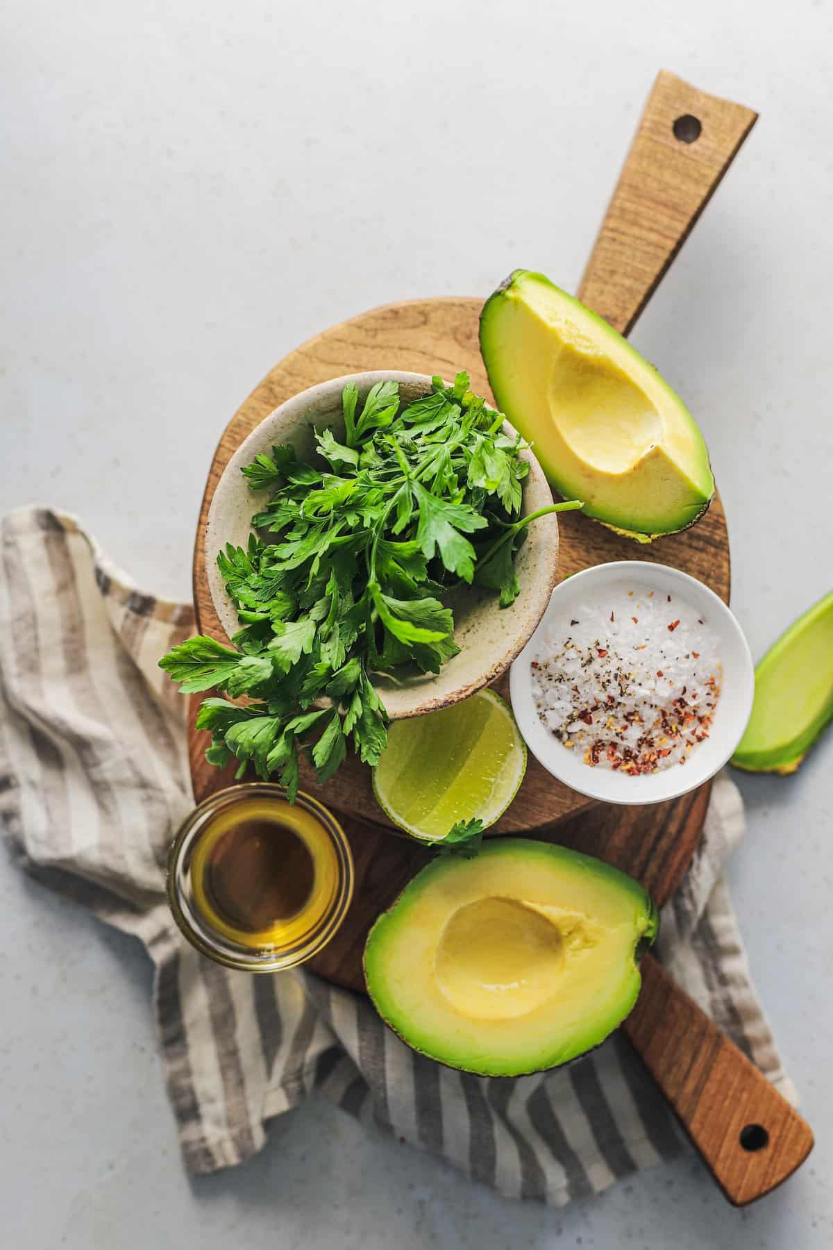 Avocados, oil, fresh cilantro and the rest of the guacamole ingredients on top of a wooden cutting board