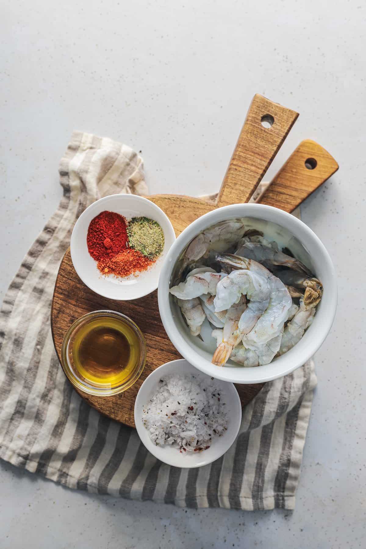 A bowl of raw shrimp beside a bowl of oil, a bowl of salt and a bowl of cajun seasoning mix