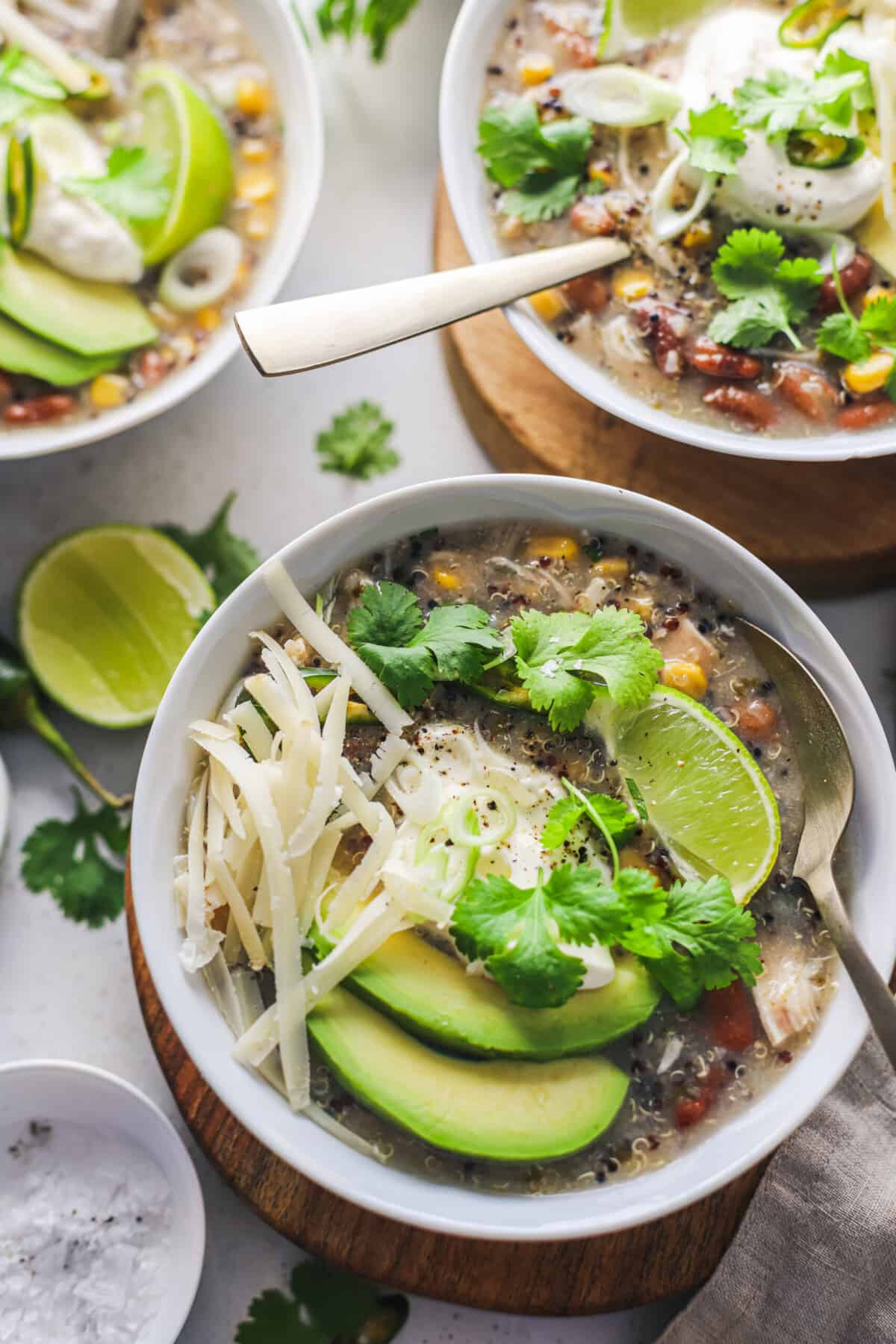 A bowl of chili garnished with avocado slices, shredded cheese, herbs and a lime wedge.