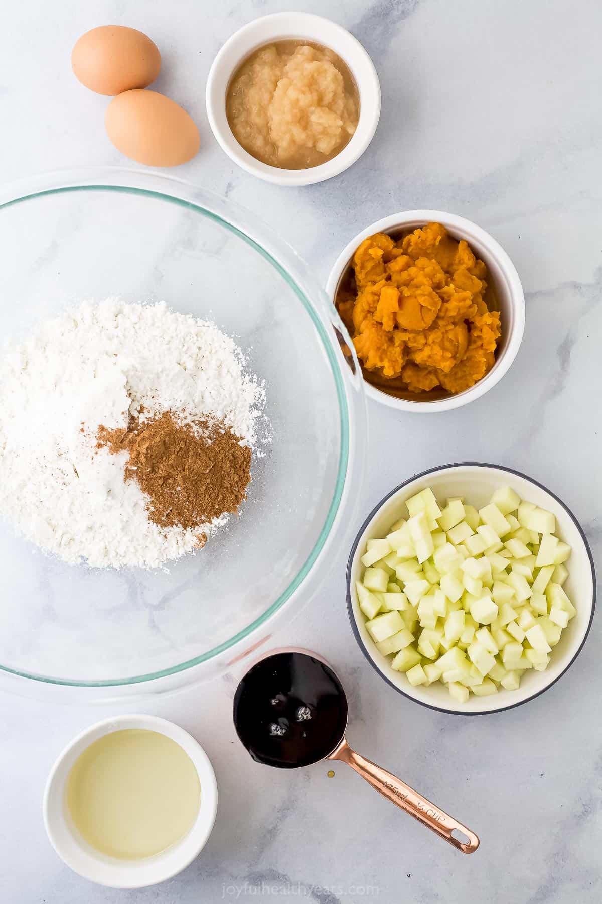 A bowl of diced apples, two eggs, applesauce and the rest of the ingredients on top of a marble counter