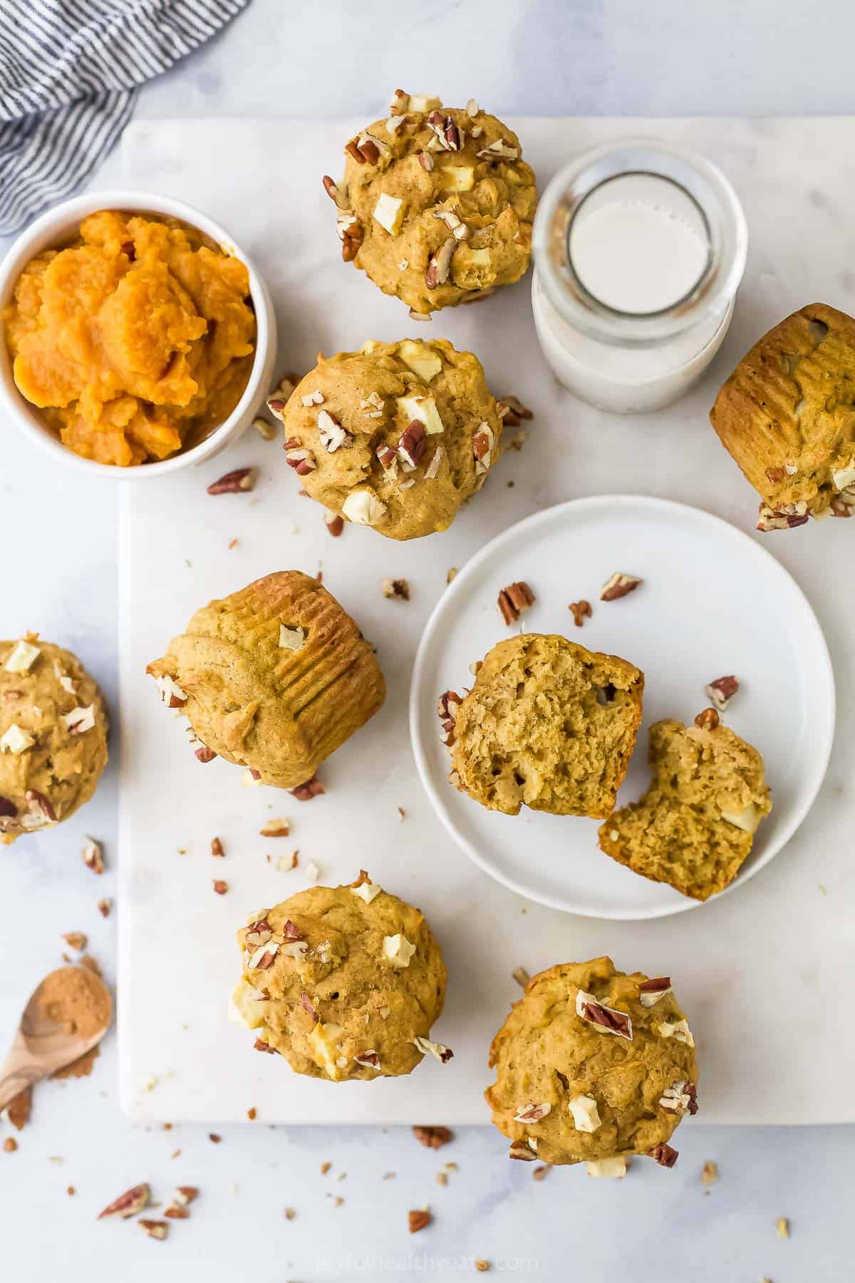 Apple pumpkin muffins scattered across a kitchen counter alongside a glass of milk and a bowl of pumpkin puree