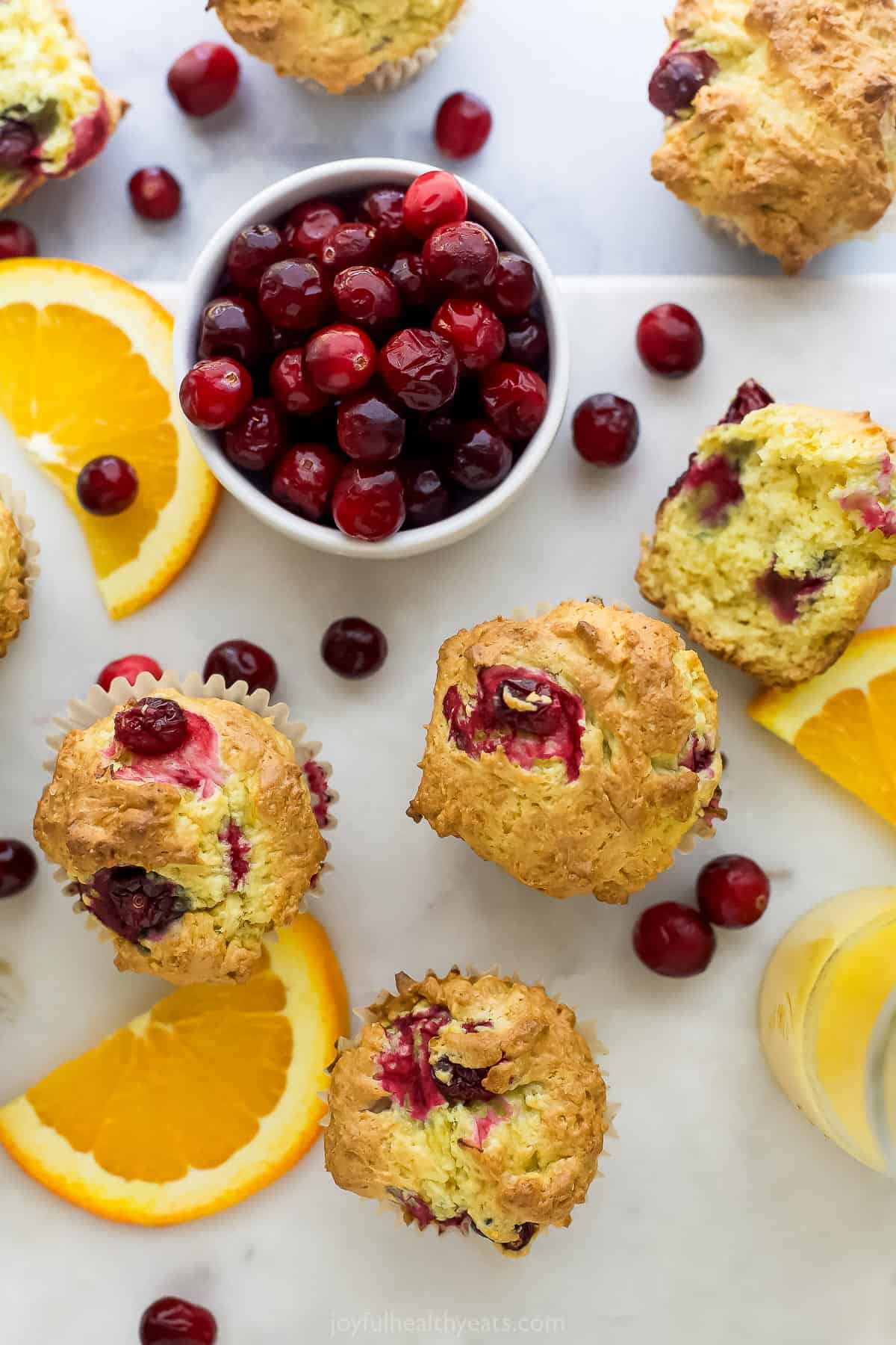 Gluten-free muffins on a countertop full of fresh fruit with one cut in half to reveal the fluffy interior