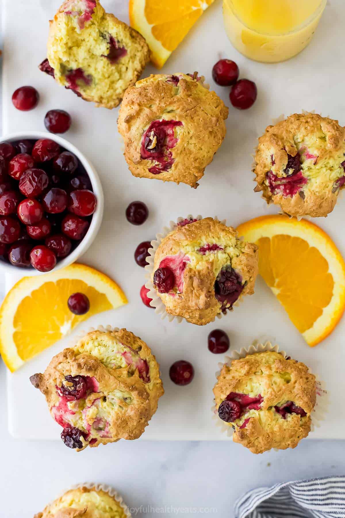 Five cranberry orange muffins on a sheet of parchment paper with orange slices and a bowl of fresh cranberries