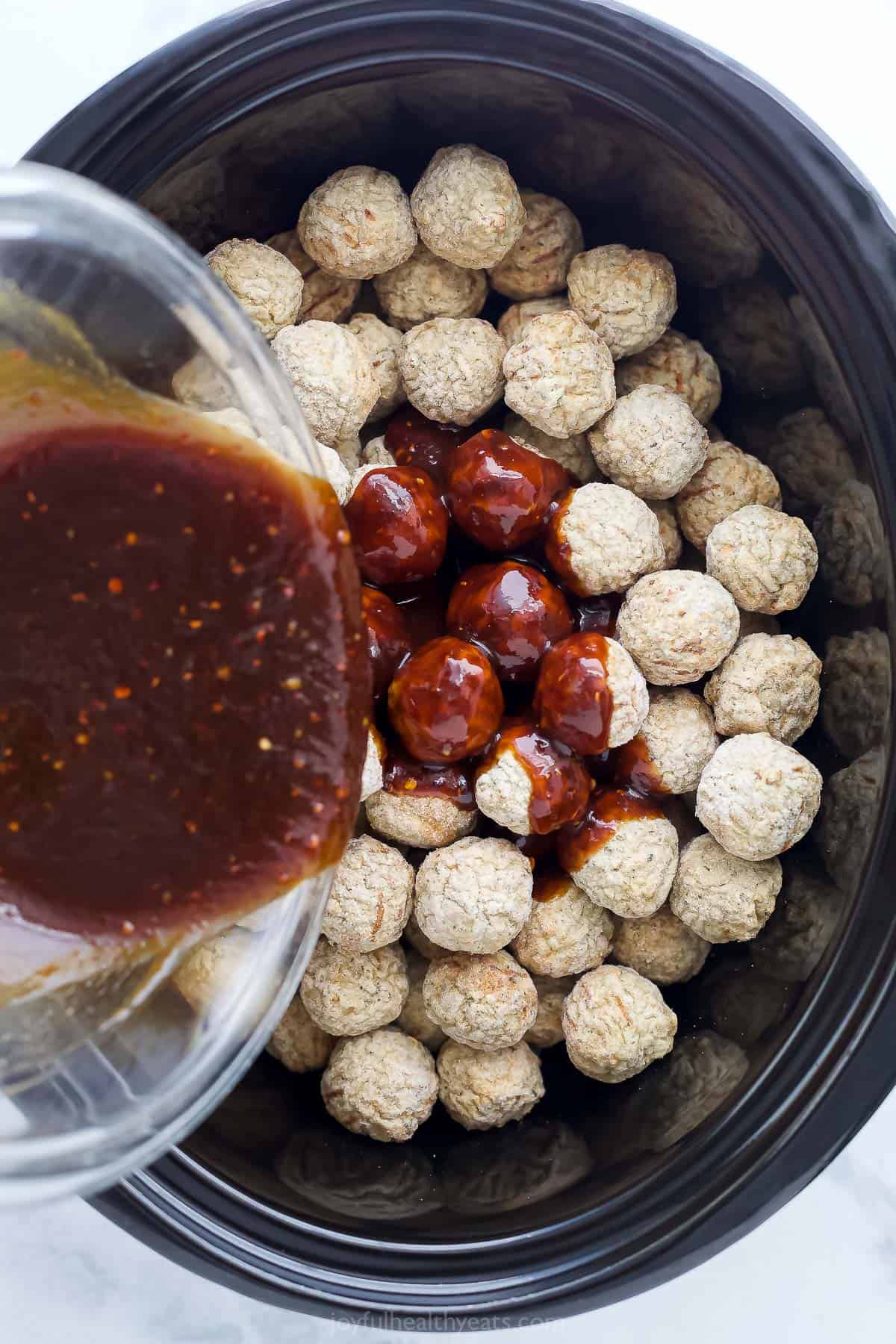 Sauce being poured into a crockpot filled with frozen meatballs