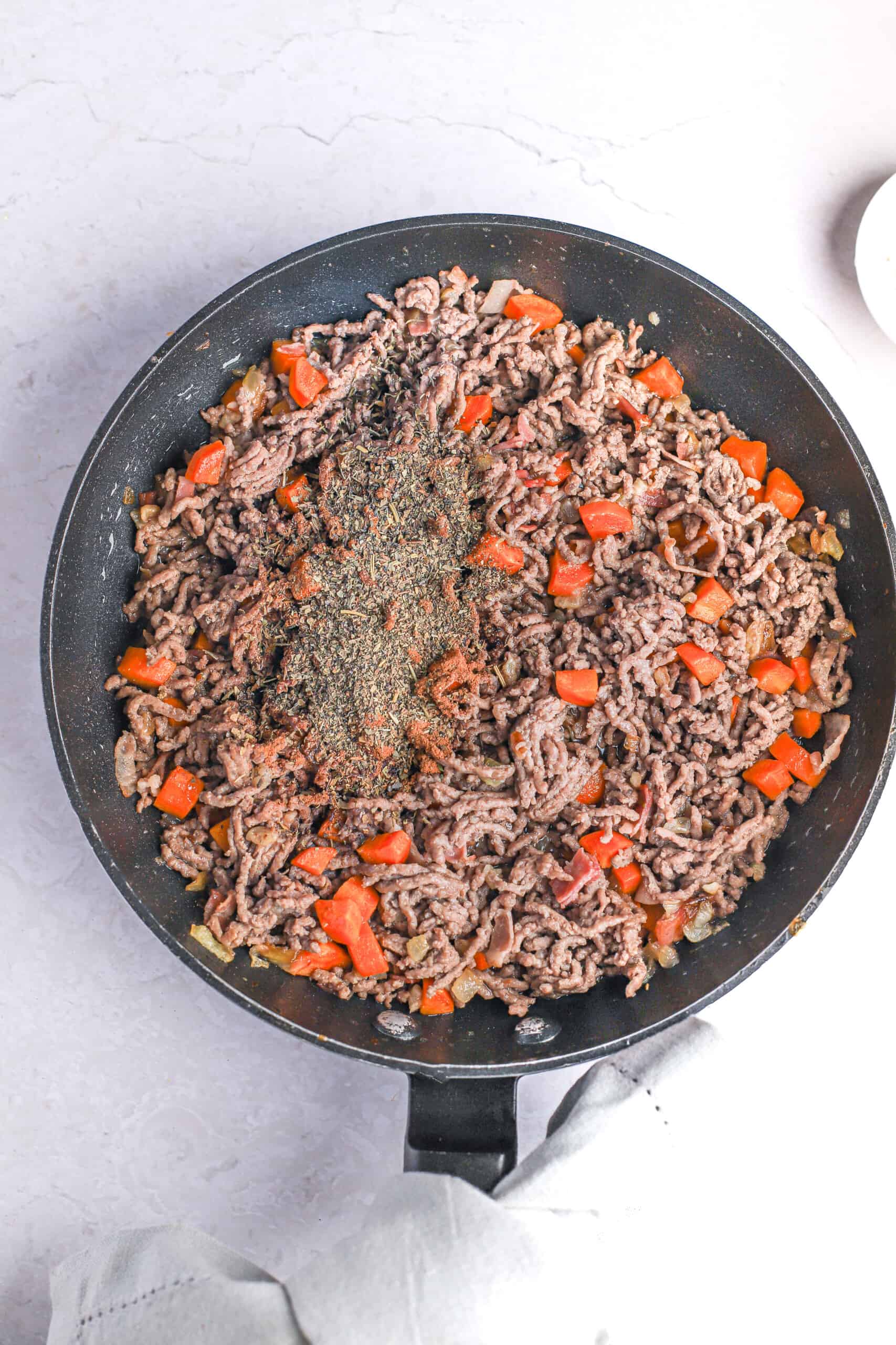 Cooked ground beef in a pan with diced onions, carrots, bacon, tomatoes and seasonings