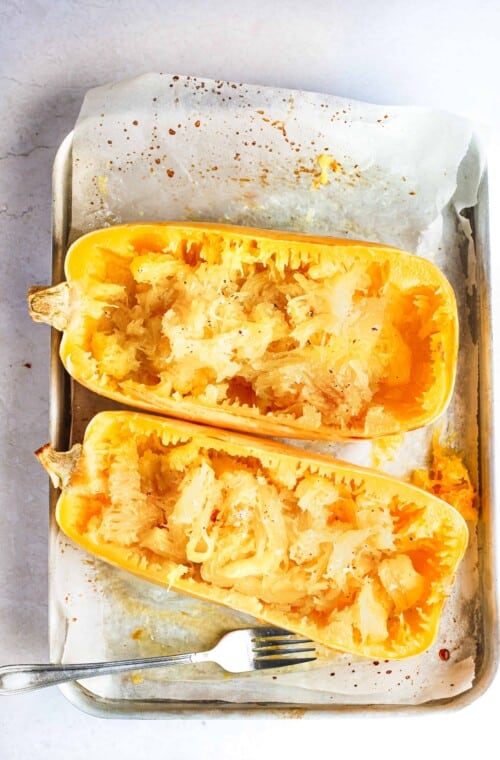 Two halves of a baked spaghetti squash on a parchment-lined pan with a fork