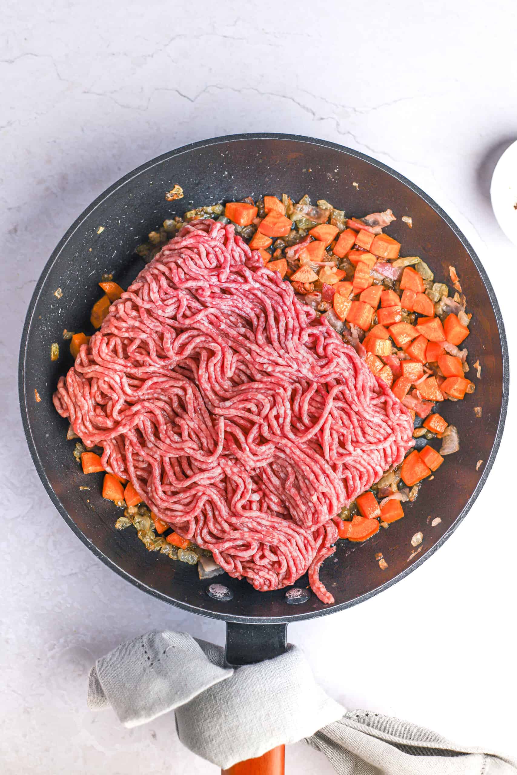 Cooked veggies and bacon in a black skillet with a pound of raw ground beef